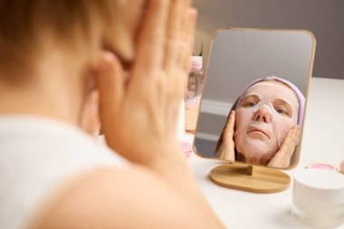 Bir kadın maske takmaya odaklanır, maskesini nazikçe yüzüne bastırır ve ahşap çerçeveli aynaya bakar. Ayar parlak, temiz bir banyo..