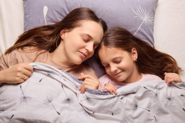 Bir anne ve kızı yumuşak bir battaniyenin altında sarılır, rahat bir yatak odasında huzurlu bir anı paylaşır, bağlarını ve rahatlıklarını vurgularlar..