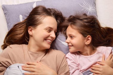Bir anne ve kızı rahat bir şekilde yataklarında uzanıyorlar, birbirlerine içtenlikle ve sevgiyle gülümsüyorlar. Huzurlu bir öğleden sonra. Mutluluk ve bağlantı dolu..