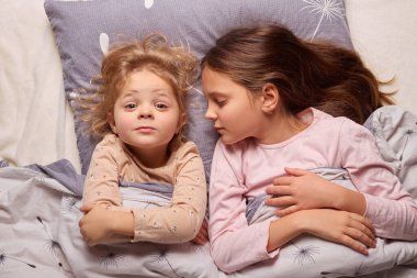 İki kız rahat battaniyelerin altında yan yana yatıyor, biri kameraya bakarken diğeri huzur içinde uyuyor. Sakin atmosfer çocukluk arkadaşlığı bağını güçlendirir..