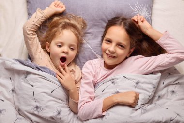 İki genç kız kardeş yataklarına kurulmuş, birlikte gülüyor ve gülüyorlar. Biri esnerken, diğeri şakacı bir şekilde esniyor. Yumuşak yatak onları sarıp sıcak bir atmosfer yaratıyor..