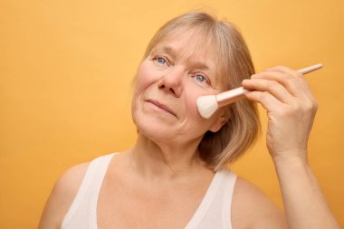 Kısa sarı saçlı bir kadın yumuşak bir makyaj fırçası kullanarak yanaklarına allık sürüyor. Parlak sarı arka plan atmosfere sıcaklık katıyor ve odaklanmış ifadesini vurguluyor..
