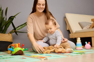 Bir anne bebeğiyle renkli ahşap oyuncaklarla dolu yumuşak bir paspasın üzerinde oyun oynamaktan hoşlanır. Konforlu odada bitkiler ve arka planda rahat bir oturma alanı bulunuyor..
