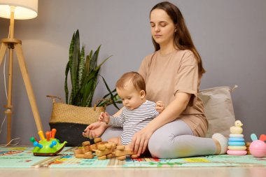 Bir anne bebeğiyle birlikte yumuşak bir mindere oturur, o da tahta blokları yığmaya odaklanır. Oyuncak ve bitkilerin yer aldığı eğlenceli bir atmosfer öğrenme deneyimini geliştirir..