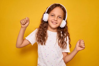 Kıvırcık saçlı bir kız, beyaz kulaklık takıp neşeli bir şekilde gülümsüyor, canlı sarı bir arka plana karşı mutlu bir şekilde dans ediyor, müziğe olan aşkını gösteriyor..
