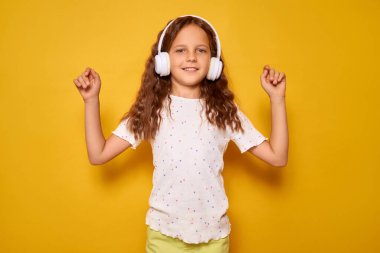 Beyaz kulaklık takan genç bir kız canlı sarı bir fonda neşeyle dans ederken gülümsüyor. Onun neşeli ifadesi ve hareketi bu canlı ortamda mutluluk saçıyor..