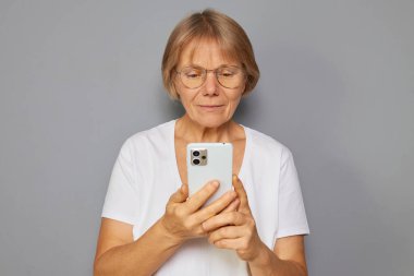 Gözlüklü ve beyaz gömlekli son sınıf öğrencisi bir kadın akıllı telefonunu odaklanmış bir ifadeyle inceliyor. Basit bir iç mekanda merak ve bağlılık gösteriyor..