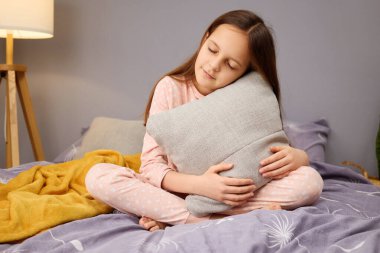 Bir kız pijamalarıyla yatağında bağdaş kurup yumuşak bir yastığa sarılıyor. Ilık aydınlatma huzurlu bir atmosfer yaratıyor, geceleri rahatlamak için mükemmel..