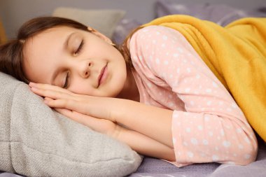 Genç bir kız, yumuşak sarı bir battaniyeyle kaplı gri bir yastığın üzerinde huzur içinde yatıyor. Puantiyeli pijama giyiyor, rahat bir yatak odasında huzurlu bir atmosfer yaratıyor..