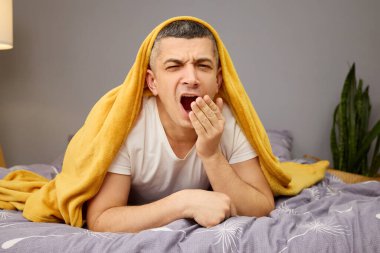 Yumuşak sarı bir battaniyenin altında yatan bir adam uyandığında esniyor. Konforlu yatak odasında hafif ışıklandırma ve biraz yeşillik var. Bu da sakin bir sabah atmosferini gösteriyor..