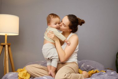 Bir anne yatakta oturup bebeğini sevgiyle kucaklar. Çocuk gülümsüyor, ve ortam sakin ve sıcak, rahat, davetkar bir yatak odası ortamında hafif ışıklandırmayla güçleniyor..