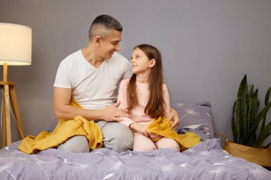 Bir baba kızıyla birlikte yatakta oturuyor. Yumuşak bir battaniyenin altında gülüyor ve birlikte vakit geçiriyorlar. Sıcak ışık sıcak odada davetkar bir atmosfer yaratıyor..