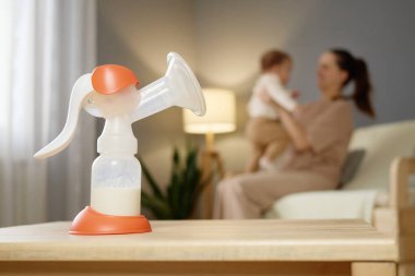 Yumuşak aydınlatılmış oturma odasındaki ahşap bir masanın üzerine belirgin bir şekilde bir göğüs pompası yerleştirilir. Arka planda, bir anne bebeğini sevgiyle kucağına alır, hassas bakım ve bağlanma anlarıyla meşgul olur..