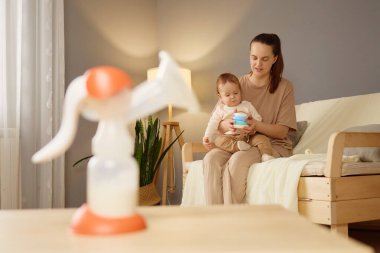 Bir anne sıcak tonlu bir oturma odasında kanepeye oturur, bebeğini nazikçe kucağına alır ve bir şişe hazırlar. Göğüs pompası yakınlardaki bir masanın üzerinde duruyor. Besleyici atmosfere katkı sağlıyor..