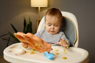 Genç bir çocuk renkli bir yemek tepsisiyle meşgul olarak yüksek bir sandalyeye rahatça oturur. Yiyecek parçaları etrafa saçılmış. Bu da sıcak ve davetkar bir odada eğlenceli bir yemek saati deneyimi olduğunu gösteriyor..