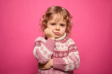 Kıvırcık saçlı genç bir çocuk rahat bir kazak giyiyor ve düşünceli bir ifadeye sahip. Çarpıcı pembe arka plan çocuğun ruh halini ön plana çıkarıyor ve atmosfere enerji katıyor..