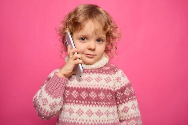 Kıvırcık saçlı bir çocuk akıllı bir telefonla sohbet ediyor, şenlik desenli, kalın örülü bir kazak giyiyor. Canlı pembe arka plan, o ana enerji katıyor..