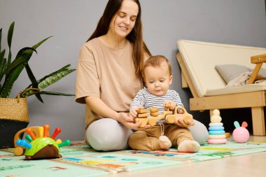 Bir anne, tahta oyuncaklar yapmakla meşgul olan küçük çocuğuyla şakalaşarak etkileşim kurar. Sıcak, davetkar bir odada çeşitli eğitici oyuncaklarla çevrili canlı bir oyun hasırının üzerinde oturuyorlar..