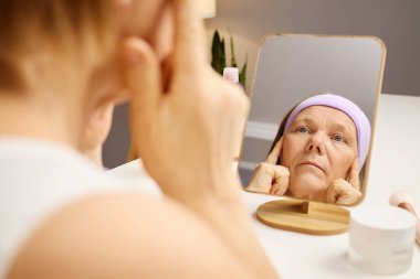Olgun bir kadın iyi aydınlatılmış bir odada cildine odaklanır. Masada otururken, aynaya bakarken nazikçe yüzüne krema sürüyor. Ortam rahatlatıcı ve kişisel..