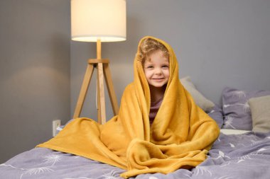 Küçük bir çocuk, sarı bir battaniyeye sarılı bir şekilde bir yatakta otururken rahat bir anın tadını çıkarır. Yakındaki bir lambadan gelen hafif ışık sıcak ve davetkar bir atmosfer yaratır..