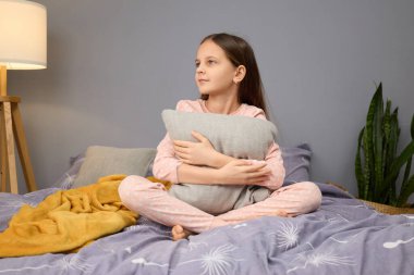 A young girl sits cross-legged on a bed, holding a soft pillow while wearing pink pajamas. The room features a warm lamp and a plant, creating a comfortable ambiance during evening time. clipart