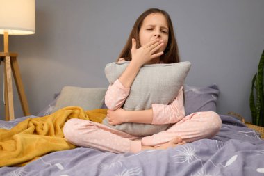 Genç bir kız, üzerinde benekler olan pijamalar giymiş, esnerken gri bir yastık tutuyor. Oda yumuşak bir şekilde aydınlatılır ve kapalı bir öğleden sonrası için dekore edilir..