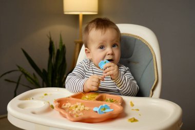 Neşeli bir bebek yüksek bir sandalyede oturur ve bölünmüş bir tabaktan yemeğin tadını çıkarır. Yemeğin kırıntıları ve kalıntıları etrafa saçılmış. Sıcak bir ortamda eğlenceli bir yemek atmosferi yaratıyor..
