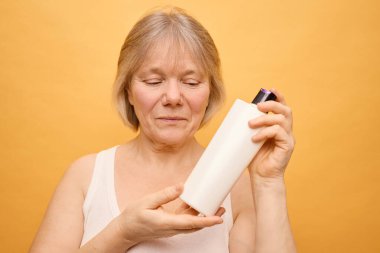 In a calm setting, a woman with short gray hair inspects a white lotion bottle, focusing on its ingredients. The cheerful yellow background adds warmth to her thoughtful expression. clipart