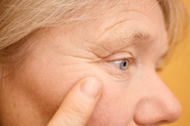 Bir kadın cildini yakından inceler, parmağını göz çevresindeki ince çizgilere dokunmak için kullanır. Aydınlatılmış bir odada, daha sağlıklı bir görünüme ve cilt bakımına odaklanıyor..