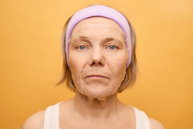 Saç bandı takmış bir kadın canlı sarı bir fonda otururken yüzüne maske takıyor. Rahatlamış ve odaklanmış görünüyor. Kişisel bakım ve cilt bakımını vurguluyor..