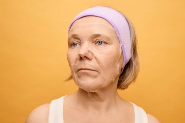 Bir kadın, cildini sulandıran bir maske takarak kendini tedavi etmeye çalışır. Rahatlamış durumda, saçları arkadan bağlanmış ve turuncu bir duvarın önünde durgun bir ifade takınıyor..