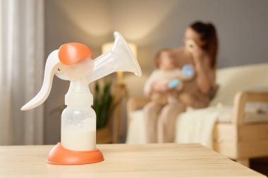 Bir anne bebeğini huzurlu bir oturma odasında tutarken bir anne tahta bir masada oturur. Yumuşak ışık, çocuğunu beslerken sıcak bir atmosfer yaratır..