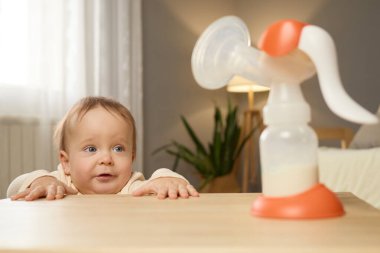 Bir bebek yakınlardaki bir göğüs pompasına dikkatle bakarak bir masaya yaslanıyor. Oda hafifçe aydınlatılıyor, arka planda bitkiler ve ışık dekoru olan rahat bir atmosfer yaratıyor..