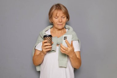 Rahat kıyafetli bir kadın bir elinde kahve fincanı tutarken diğer elinde akıllı telefonunu kontrol eder ve yumuşak gri bir arka planda durur. İfadesi ilgi ve bağlılık gösteriyor..