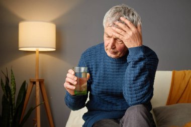 Yaşlı bir adam sıcak bir oturma odasında bir kanepede oturuyor, elinde bir bardak içecekle. Bir lamba etrafındaki boşluğu hafifçe aydınlatırken o derin düşüncelerin içinde belirir..