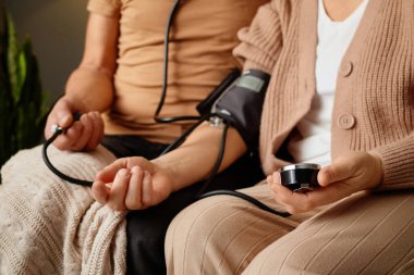 Bir insan rahat bir odada rahatça otururken kan basıncını cihazla ölçüyor. Partnerleri süreci gözlemleyerek sağlık denetimi için destekleyici bir atmosfer yaratıyor.