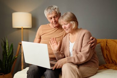 Üst düzey bir çift dizüstü bilgisayarla görüntülü konuşma yapmaktan hoşlanıyor. Sıcak ışıklandırma ve rahat dekor ile çevrili bir kanepeye yakın otururlar. Konuşmaları sırasında gülümser ve el sallarlar..