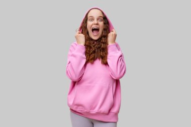 Kıvırcık saçlı genç bir kadın heyecanla gülümsüyor ve pembe kapüşonlu bir poz veriyor. Neşeli ifadesi, basit bir ev ortamında heyecan ve mutluluk yayıyor..