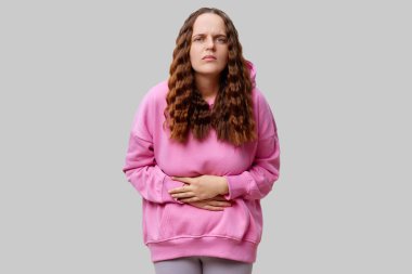 A young woman with long, wavy hair stands with a pained expression, clutching her stomach. She wears a pink hoodie and grey pants against a plain background, conveying discomfort and unease. clipart