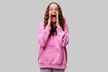 Uzun, dalgalı saçlı genç bir kadın pembe bir kapüşonlu giyer ve ağzını dramatik bir şekilde açıp ellerini yüzüne kaldırarak heyecan gösterir. İfadesi saf neşe veriyor..