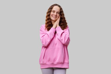 Kıvırcık saçlı neşeli genç bir kadın pembe bir kazak giyer ve elleri yanaklarında poz verir. Tarafsız bir ortamda sıcaklığını ve mutluluğunu ifade eder..