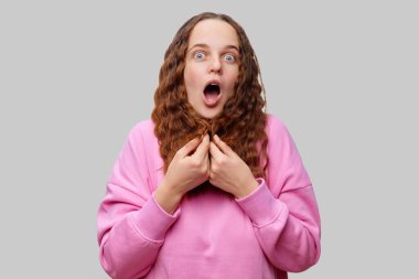 Kıvırcık saçlı genç bir kadın şok edici bir ifade sergiler. Pembe bir kazak giyiyor ve içeride sürpriz bir habere ya da beklenmedik bir olaya tepki veriyor..