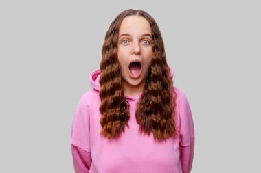 Uzun dalgalı saçlı bir kadın, ağzı açık bir şekilde sürpriz yapıyor. Pembe bir kapüşonlu giyiyor. Tarafsız gri bir arka plana yaslanmış, çarpıcı bir kontrast oluşturuyor..