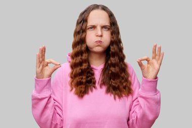 Dalgalı saçlı bir kadın pembe bir kapüşonlu giyer ve yanaklarını şişirerek hayal kırıklığını ifade eder. Sakin bir jest yaparak ellerini tutuyor. Sinirli ifadesine zıt düşüyor..