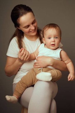 Bir anne, rahat giyimli bebeğini kucağında tutarken gülümser ve sakin bir ortamda hassas bir bağ gösterir. Atmosfer sıcak ve samimi..