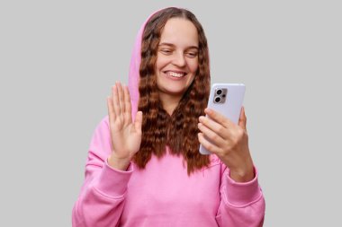 Pembe kapüşonlu uzun, dalgalı saçlı neşeli genç bir kadın bir video çağrısına katılırken geniş bir yelpazede gülümsüyor, basit bir arka planda coşkuyla ekrana el sallıyor..