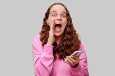 Kıvırcık saçlı genç bir kadın akıllı telefonunu tutarken neşeyle bağırıyor. Parlak pembe bir kapüşonlu giyiyordu ve yüzünde mutluluk ya da iyi haberi simgeleyen heyecanlı bir ifade vardı..