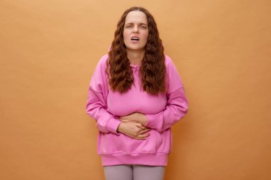 Bir kadın acı dolu bir ifadeyle ayakta duruyor, karnını sıkıyor. Pembe bir kapüşonlu ve açık renk pantolon giyiyor, canlı bir ortamda duygusal ve fiziksel rahatsızlığını gösteriyor..