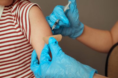 Mavi eldivenli bir sağlık görevlisi aşı yapmaya hazırlanırken küçük bir çocuk klinikte rahat bir şekilde oturuyor. Çocuk sakin bir tavır sergiliyor, prosedüre hazır..