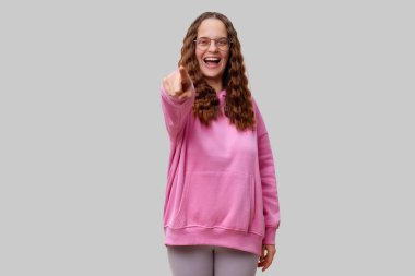 Yumuşak pembe kapüşonlu neşeli genç bir kadın coşkusunu geniş bir şekilde gülümseyerek ve neşeyle ileriye doğru göstererek dostça bir atmosfer yaratarak gösteriyor..